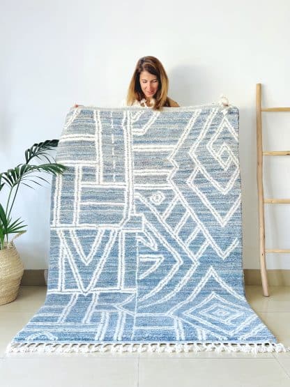 Woman holding a handmade, Moroccan rug with white line designs on a mixed blue background.