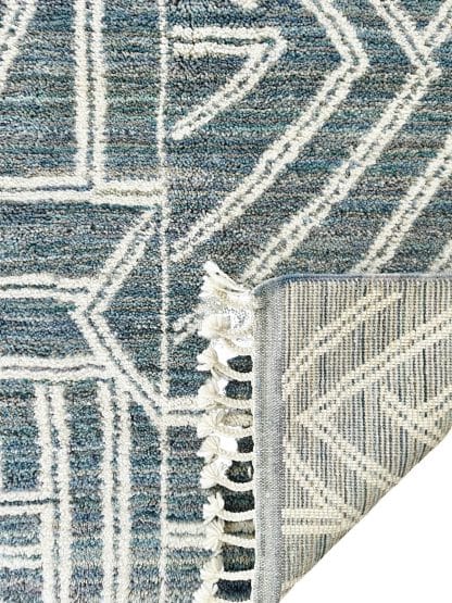 Close up of a handmade, Moroccan rug with white line designs on a mixed blue background. One corner is folded up.