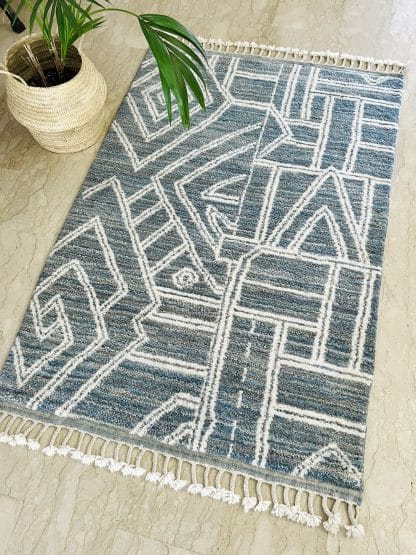 A handmade, Moroccan rug with white line designs on a mixed blue background in a room with marble floors and a plant.