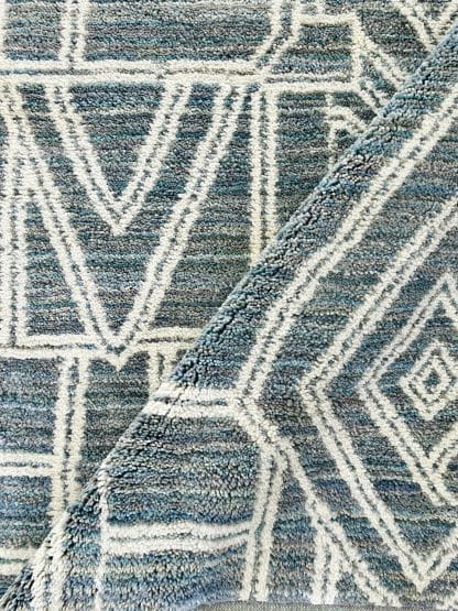 Close up shot of a folded, handmade, Moroccan rug with white line designs on a mixed blue background.