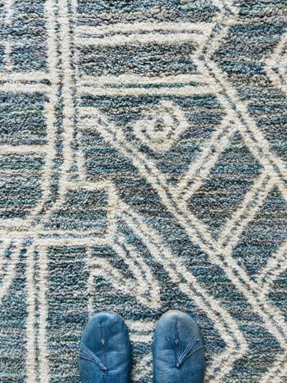 Close up of a handmade, Moroccan rug with white line designs on a mixed blue background. There are feet at the bottom of the image in blue slippers.