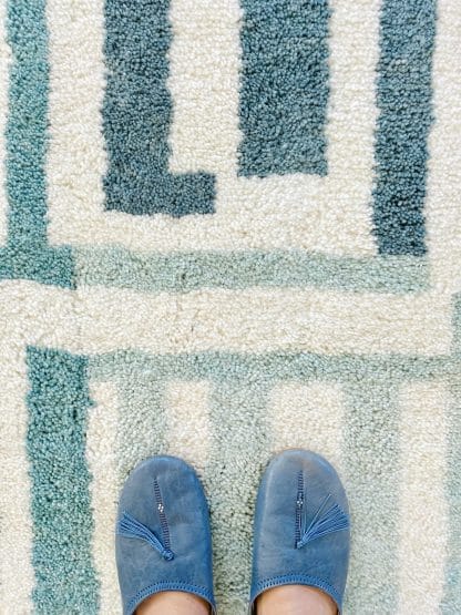 Close up of a Beni Ourain Moroccan Rug with a blue and white maze design. A person is standing on the rug but just their feet are visible at the bottom of the image.