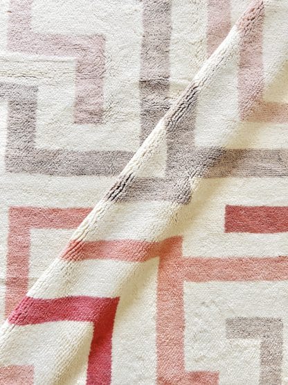 Close up of a Beni Ourain Moroccan Rug with a bold cream, beige and pink maze design. There is a diagonal fold in the rug.