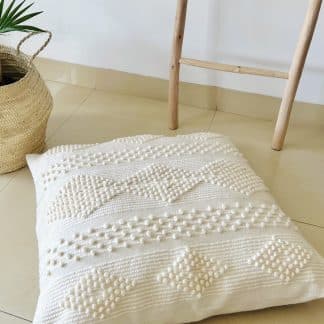 Moroccan floor pillow made of off-white wool with little woollen balls arranged in geometric designs on a marble floor. There is a plant and part of a wooden ladder in the shot.