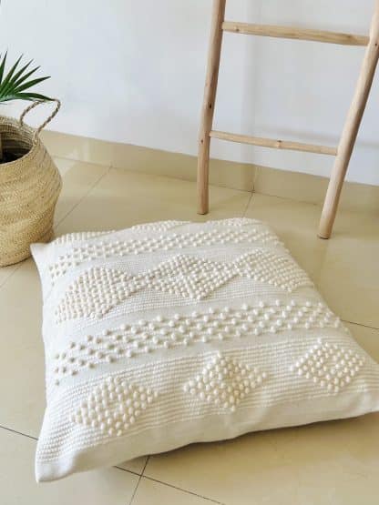 Moroccan floor pillow made of off-white wool with little woollen balls arranged in geometric designs on a marble floor. There is a plant and part of a wooden ladder in the shot.