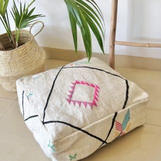 Off-white Moroccan floor pouf handmade from a traditional flat weave kilim with multicoloured Berber designs on a marble floor with a plant and wooden ladder in the background.