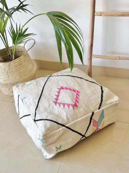 Off-white Moroccan floor pouf handmade from a traditional flat weave kilim with multicoloured Berber designs on a marble floor with a plant and wooden ladder in the background.
