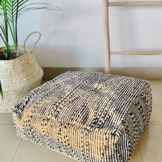 Black and white Moroccan floor pouf handmade from a traditional zanafi kilim with raised designs on a marble floor with a plant and wooden ladder in the background.