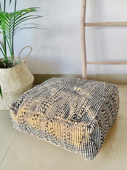 Black and white Moroccan floor pouf handmade from a traditional zanafi kilim with raised designs on a marble floor with a plant and wooden ladder in the background.