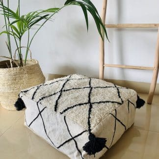 Black and white Moroccan floor pouf handmade from a traditional flat weave kilim and a beni ourain rug with black diamond designs and black tassels on the corners on a marble floor with a plant and wooden ladder in the background.