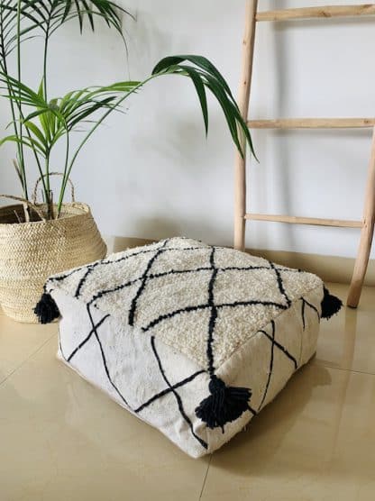 Black and white Moroccan floor pouf handmade from a traditional flat weave kilim and a beni ourain rug with black diamond designs and black tassels on the corners on a marble floor with a plant and wooden ladder in the background.