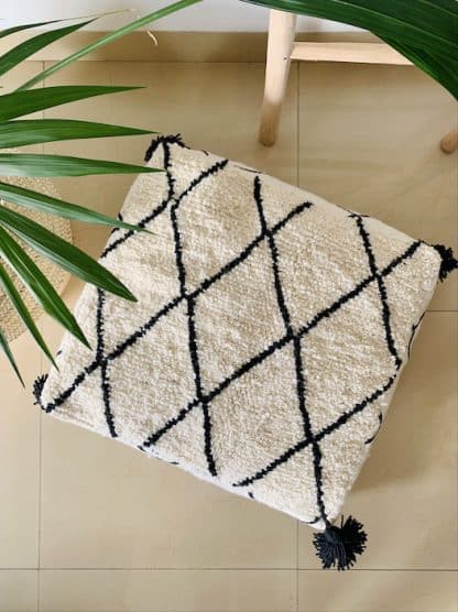 Aerial shot of a black and white Moroccan floor pouf handmade from a traditional flat weave kilim and a beni ourain rug with black diamond designs and black tassels on the corners on a marble floor with a plant and wooden ladder.