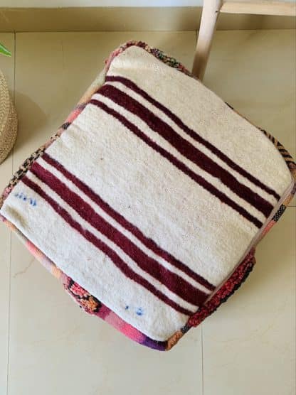 Aerial shot of an upside down colourful Moroccan floor pouf handmade from a vintage flat weave kilim with multicoloured Berber designs on a marble floor with a plant and wooden ladder in the background.