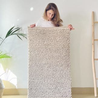 Handmade Moroccan rug Tapis de Boules in beige held by a woman with a plant and a wood ladder in frame on either side.