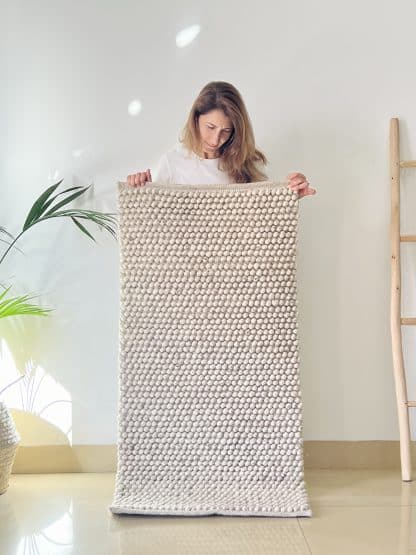 Handmade Moroccan rug Tapis de Boules in beige held by a woman with a plant and a wood ladder in frame on either side.