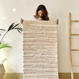 Women holding a Tapis de Boules Ivory Wave rug with a plant and a wooden ladder on either side.