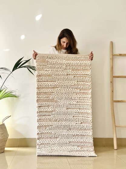 Women holding a Tapis de Boules Ivory Wave rug with a plant and a wooden ladder on either side.