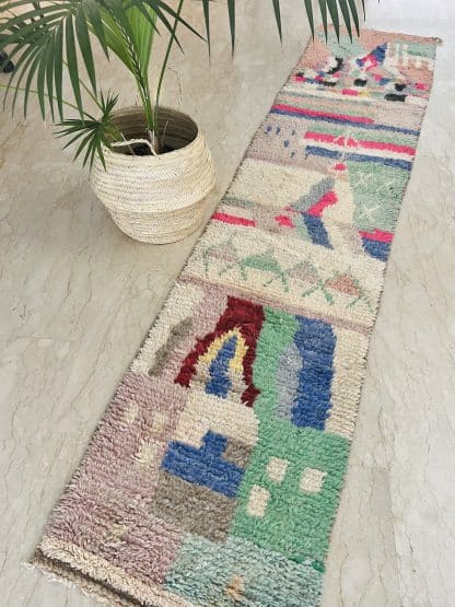 Aerial shot of a Moroccan rug, Azilal Runner - Tropical. A hand-knotted, runner rug with a unique, colourful design. It is laid on a marble floor with a palm plant nearby.