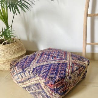 Moroccan Floor Pouf handmade from a vintage rug. It features geometric Berber designs pastel shades of pink, purple and beige. Pictured on a marble floor with a wooden ladder and palm plant in the shot.
