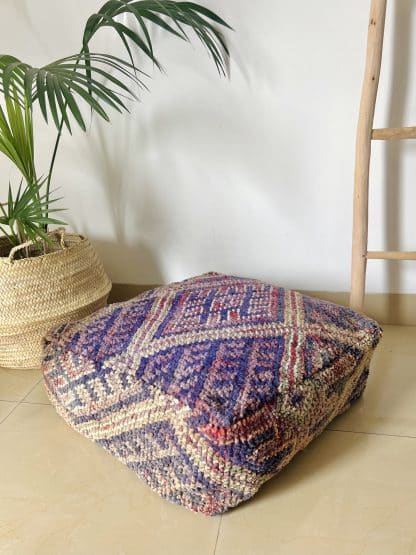 Moroccan Floor Pouf handmade from a vintage rug. It features geometric Berber designs pastel shades of pink, purple and beige. Pictured on a marble floor with a wooden ladder and palm plant in the shot.