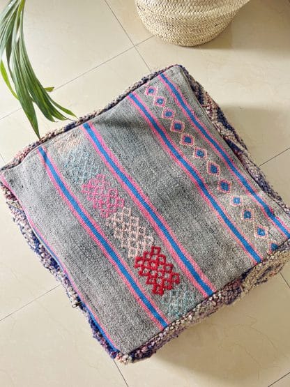 Upside down Moroccan Floor Pouf handmade from a vintage rug. It features geometric Berber designs pastel shades of pink, purple and beige. Pictured on a marble floor with a palm plant in the shot.