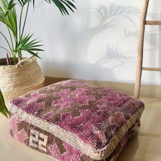 Multicoloured Moroccan Floor Pouf handmade from a vintage rug. It features geometric Berber designs in shades of pink and brown. Pictured on a marble floor with a palm plant and wooden ladder.