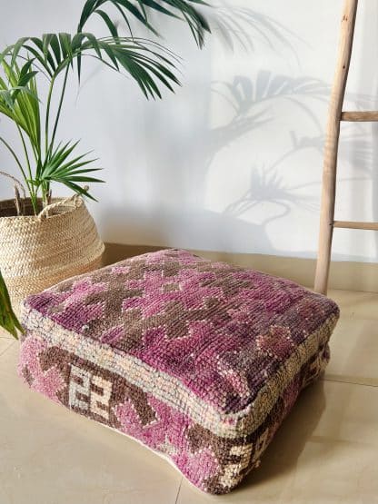 Multicoloured Moroccan Floor Pouf handmade from a vintage rug. It features geometric Berber designs in shades of pink and brown. Pictured on a marble floor with a palm plant and wooden ladder.
