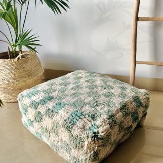 Moroccan floor pouf handmade from a vintage medium pile rug. It features a modern checkered design in off-white and pastel teal. Pictured on a marble floor with a wooden ladder and palm plant in the background.