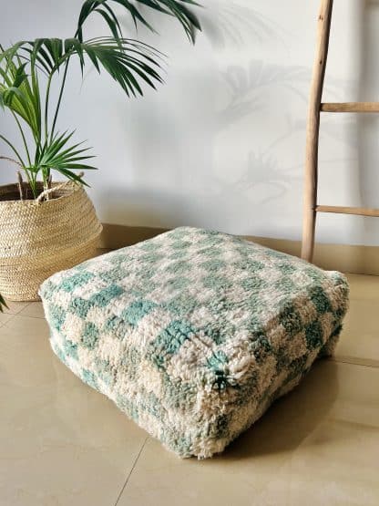 Moroccan floor pouf handmade from a vintage medium pile rug. It features a modern checkered design in off-white and pastel teal. Pictured on a marble floor with a wooden ladder and palm plant in the background.