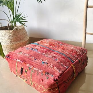 Moroccan floor pouf handmade from a vintage flat weaved kilim. It features a light red background with multicoloured simple geometric Berber designs. Pictured on a marble floor with a plant and wooden ladder in the background.