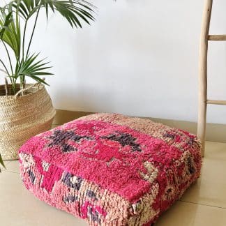 Moroccan floor pouf handmade from a vintage low pile rug. It features geometric Berber designs in shades of hot pink, charcoal grey and beige. Pictured on a marble floor with a plant and wooden ladder in the background.
