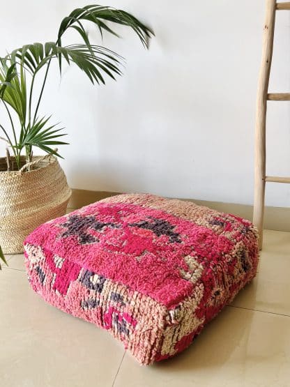 Moroccan floor pouf handmade from a vintage low pile rug. It features geometric Berber designs in shades of hot pink, charcoal grey and beige. Pictured on a marble floor with a plant and wooden ladder in the background.