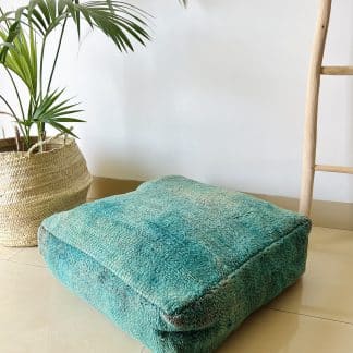 Moroccan floor pouf handmade from a vintage low pile rug. It features different shades of blue and teal. Pictured on a marble floor with a plant and wooden ladder in the background.