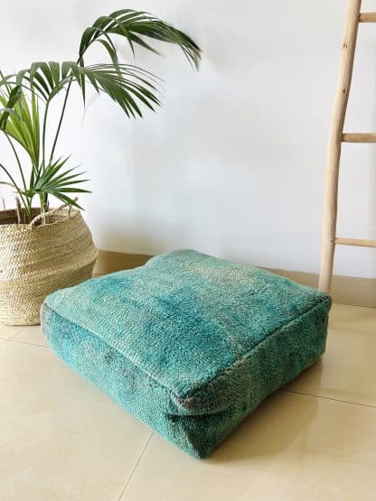 Moroccan floor pouf handmade from a vintage low pile rug. It features different shades of blue and teal. Pictured on a marble floor with a plant and wooden ladder in the background.