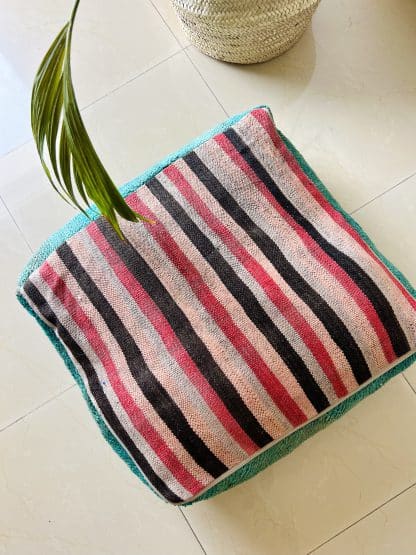 Upside down Moroccan floor pouf handmade from a vintage low pile rug. It features different shades of blue and teal. Pictured on a marble floor with a plant.