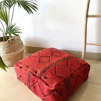 Moroccan floor pouf handmade from a vintage flat weaved kilim. It features a dark red background with simple black geometric Berber designs. Pictured on a marble floor with a plant and wooden ladder in the background.