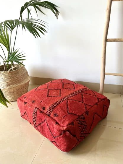 Moroccan floor pouf handmade from a vintage flat weaved kilim. It features a dark red background with simple black geometric Berber designs. Pictured on a marble floor with a plant and wooden ladder in the background.