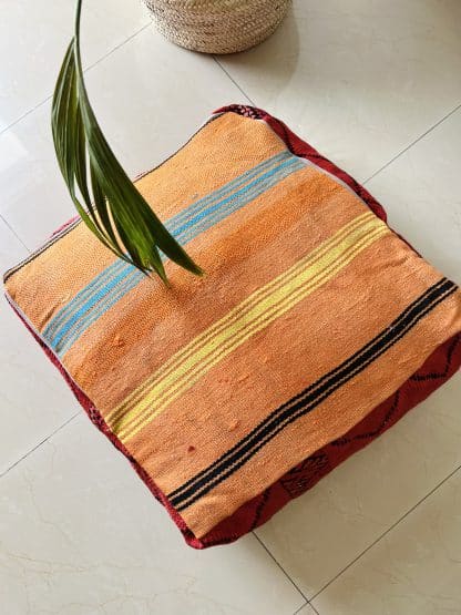 Upside down Moroccan floor pouf handmade from a vintage flat weaved kilim. It features a dark red background with simple black geometric Berber designs. Pictured on a marble floor with a plant.