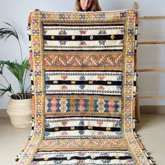 Woman holding an off-white, black and orange Moroccan rug with tufted designs in shades of pink, blue, and yellow. Pictured with a palm plant and a wooden ladder.