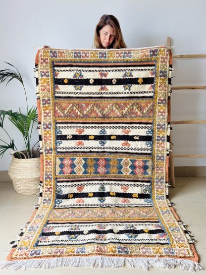 Woman holding an off-white, black and orange Moroccan rug with tufted designs in shades of pink, blue, and yellow. Pictured with a palm plant and a wooden ladder.