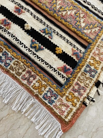 Close up shot of the corner of an off-white, black and orange Moroccan rug with tufted designs in shades of pink, blue, and yellow.