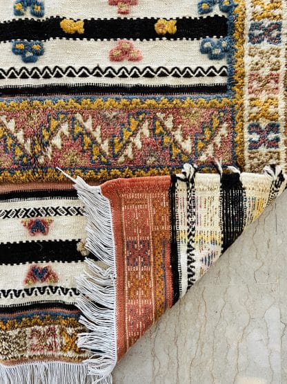 Close up shot of the folded corner of an off-white, black and orange Moroccan rug with tufted designs in shades of pink, blue, and yellow.