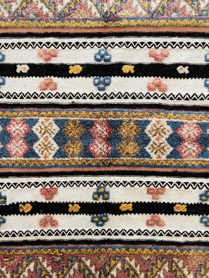 Close up shot of an off-white, black and orange Moroccan rug with tufted designs in shades of pink, blue, and yellow.