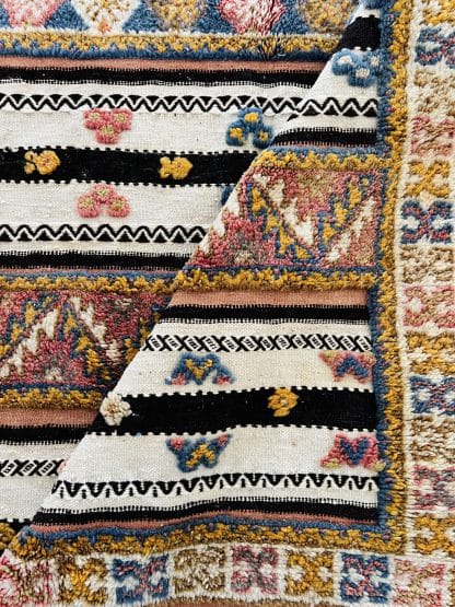 Close up shot of the a fold in the centre of an off-white, black and orange Moroccan rug with tufted designs in shades of pink, blue, and yellow.