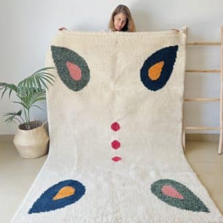 Woman holding a Beni Ourain Moroccan Rug created in collaboration with White Otter Design Co. It features a cream base with green and blue petals, and red dots.