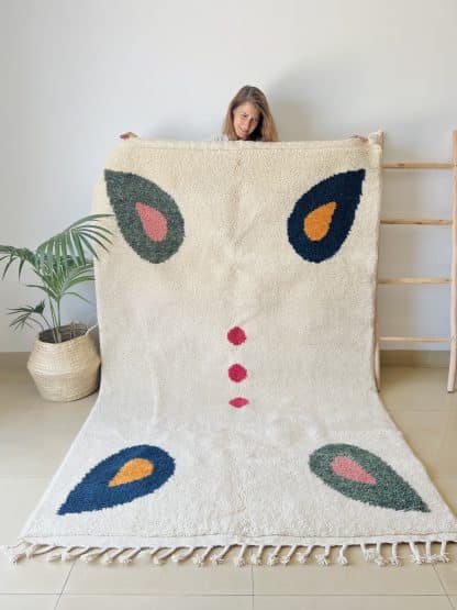 Woman holding a Beni Ourain Moroccan Rug created in collaboration with White Otter Design Co. It features a cream base with green and blue petals, and red dots.