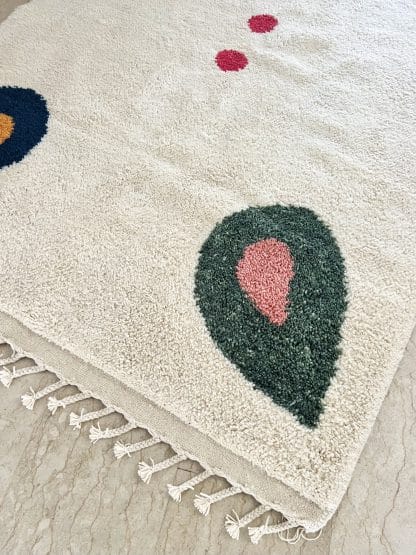 Close up of the corner of a Beni Ourain Moroccan Rug created in collaboration with White Otter Design Co. It features a cream base with green and blue petals, and red dots.