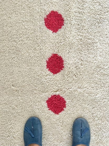 Close up of the centre red dots of a a Beni Ourain Moroccan Rug created in collaboration with White Otter Design Co. It features a cream base with green and blue petals, and red dots. There is a person's feet in blue Moroccan slippers in the shot.