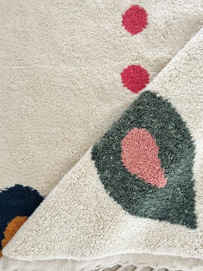 Close up of a fold in the centre of a Beni Ourain Moroccan Rug created in collaboration with White Otter Design Co. It features a cream base with green and blue petals, and red dots.