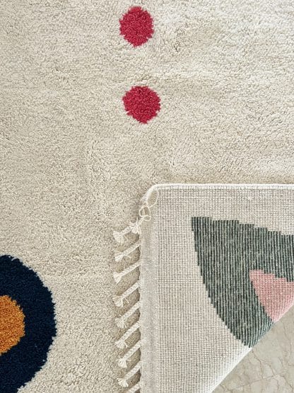 Close up of the folded corner of a Beni Ourain Moroccan Rug created in collaboration with White Otter Design Co. It features a cream base with green and blue petals, and red dots.